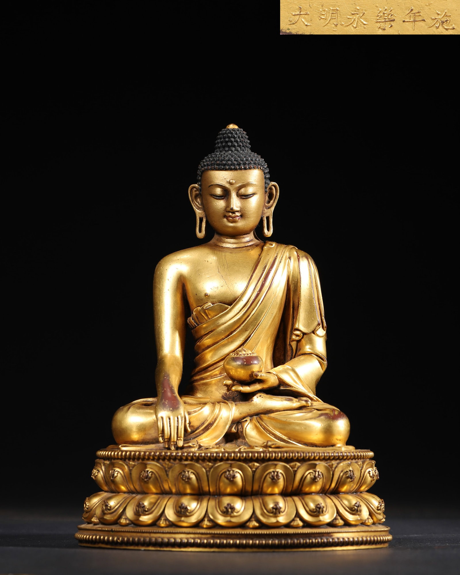 A GILT-BRONZE FIGURE OF MEDICINE BUDDHA: A gilt-bronze figure of Medicine Buddha seated on a lotus base, the right hand lowered and the left holding a medicine bowl. The base bears a crossed-vajra motif. L. 13.5 cm; W. 12.5 cm; Ht. 19 cm; Wt