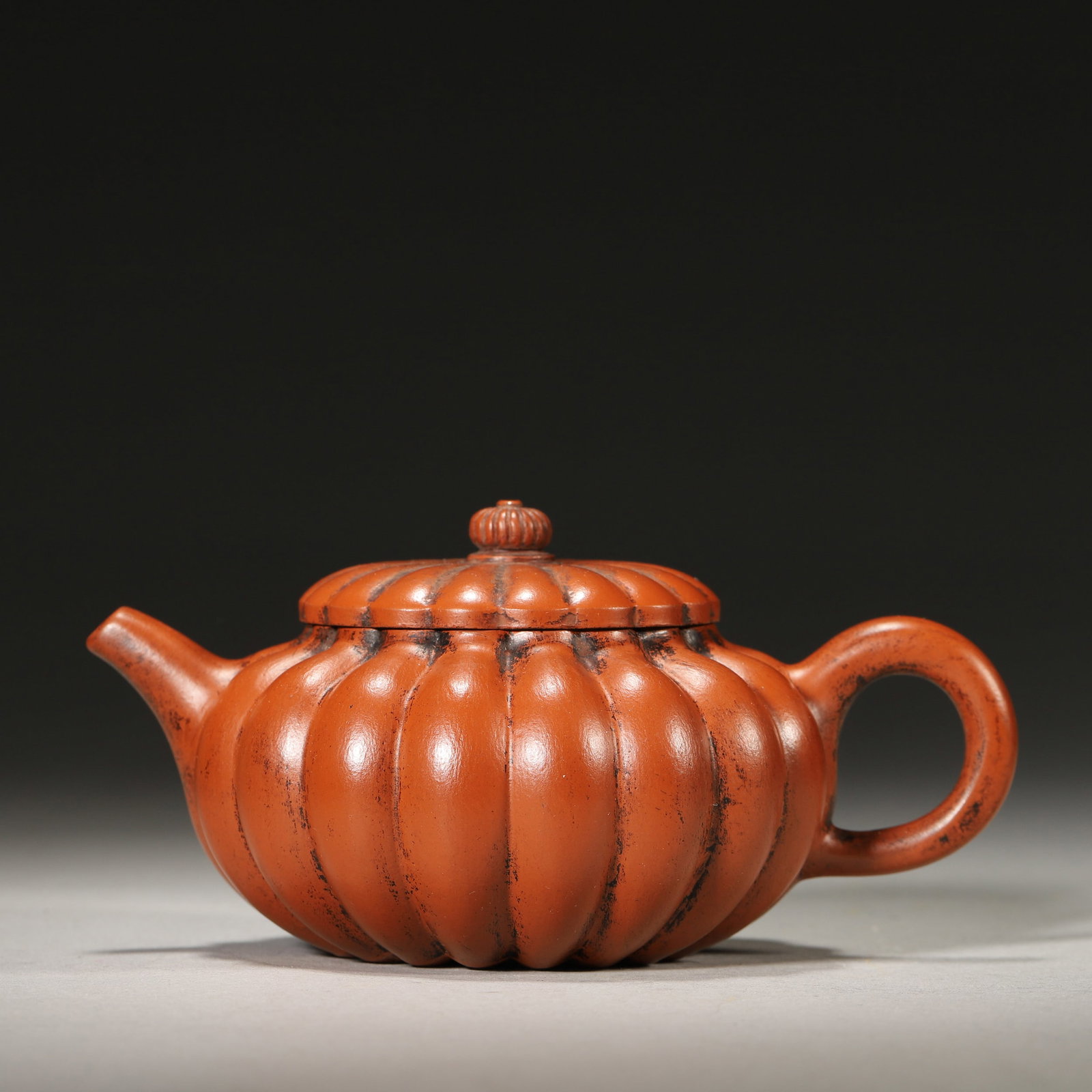 A ZISHA CHRYSANTHEMUM-FORM TEAPOT: A red-clay zisha teapot with chrysanthemum-ribbed body and cover and a flower-core-form finial. The base bears a mark. L. 12.5 cm; W. 8.5 cm; Ht. 6.5 cm; Wt. 134 g.