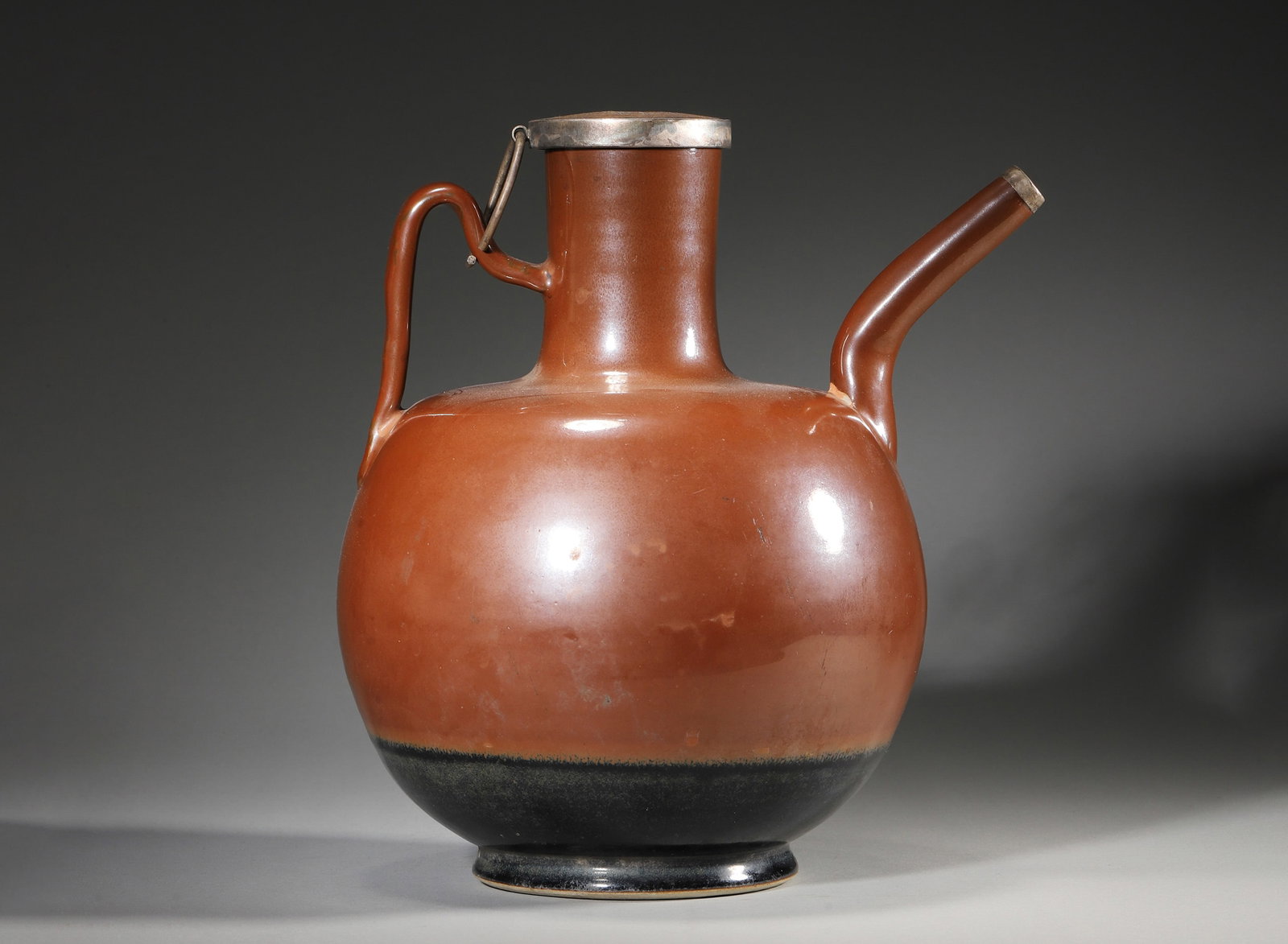 A BROWN-GLAZED PORCELAIN EWER WITH METAL COVER: A porcelain ewer covered in a reddish-brown glaze with a black-glazed lower section, fitted with a spout and handle. The metal cover is engraved with birds and flowers and bears an inscription