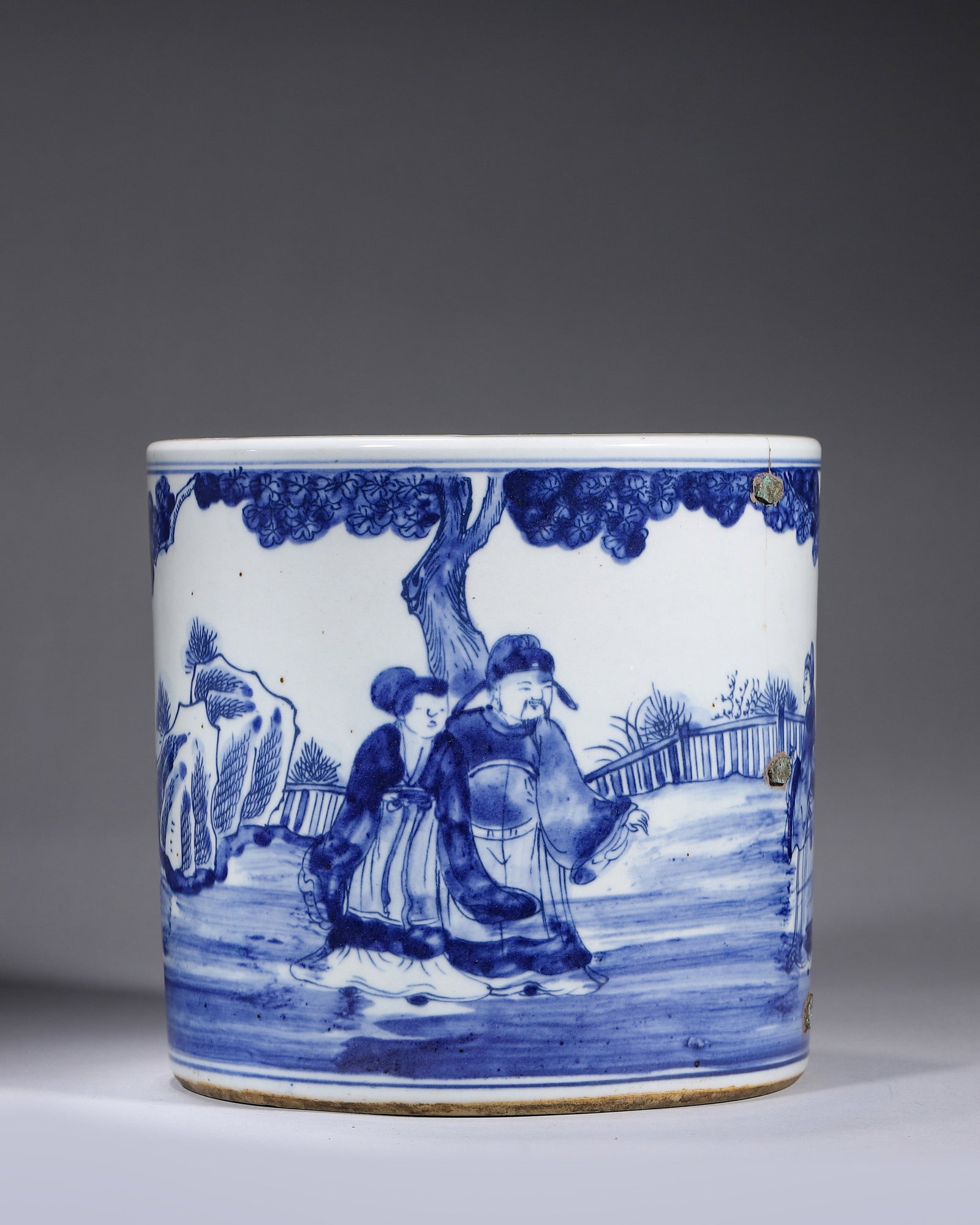 A BLUE-AND-WHITE 'SCHOLARS IN A GARDEN' BRUSH POT: A cylindrical blue-and-white porcelain brush pot painted with scholars and attendants in a garden with pines and rockwork. Diam. 20 cm; Ht. 19 cm. Condition: Metal staples from an old repair.