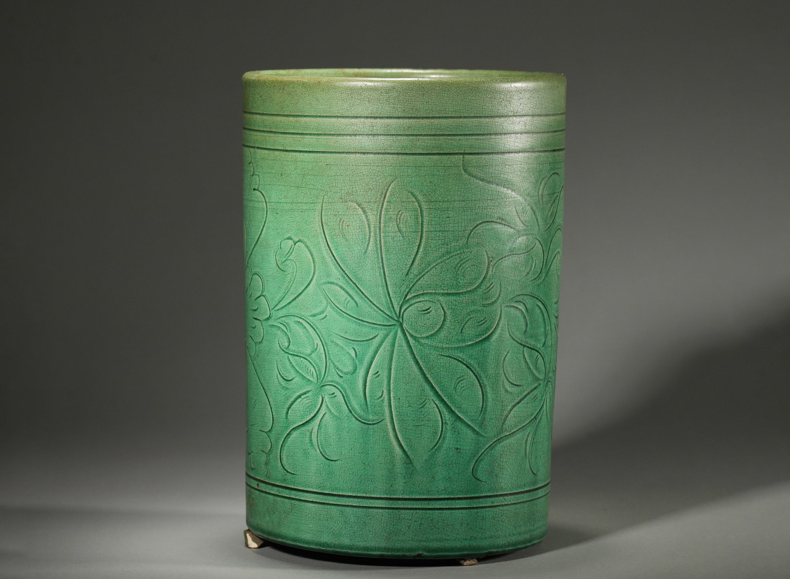 A GREEN-GLAZED CARVED FLORAL CYLINDRICAL CENSER: A cylindrical porcelain censer covered in a green crackle glaze, carved with scrolling flowers and horizontal bands, on three feet. Ht. 21 cm.