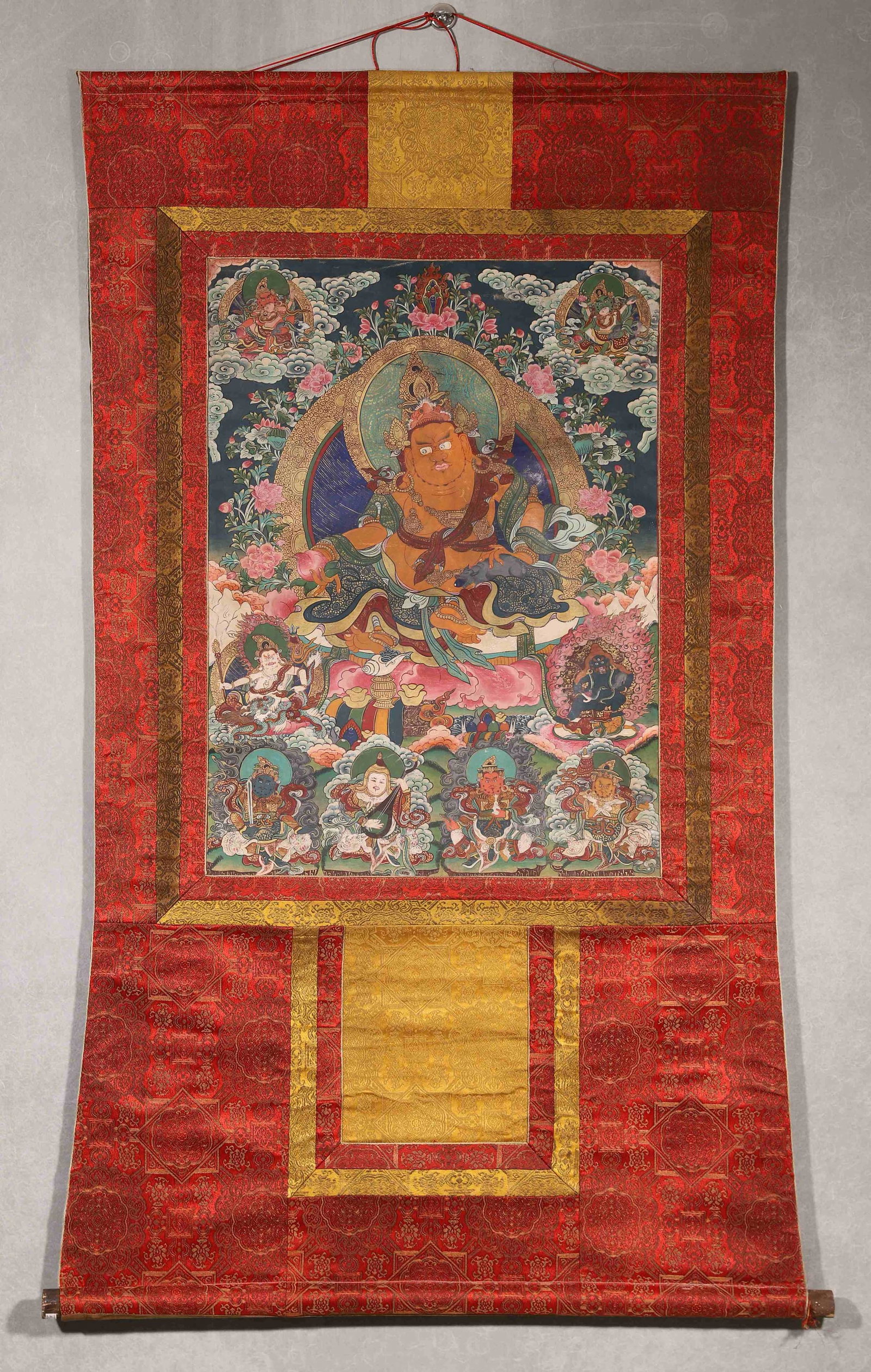 A THANGKA OF YELLOW JAMBHALA: A mineral-pigment thangka depicting Yellow Jambhala holding a mongoose, surrounded by wealth deities and protectors, mounted in red-ground brocade with wooden rollers. The reverse bears cinnabar