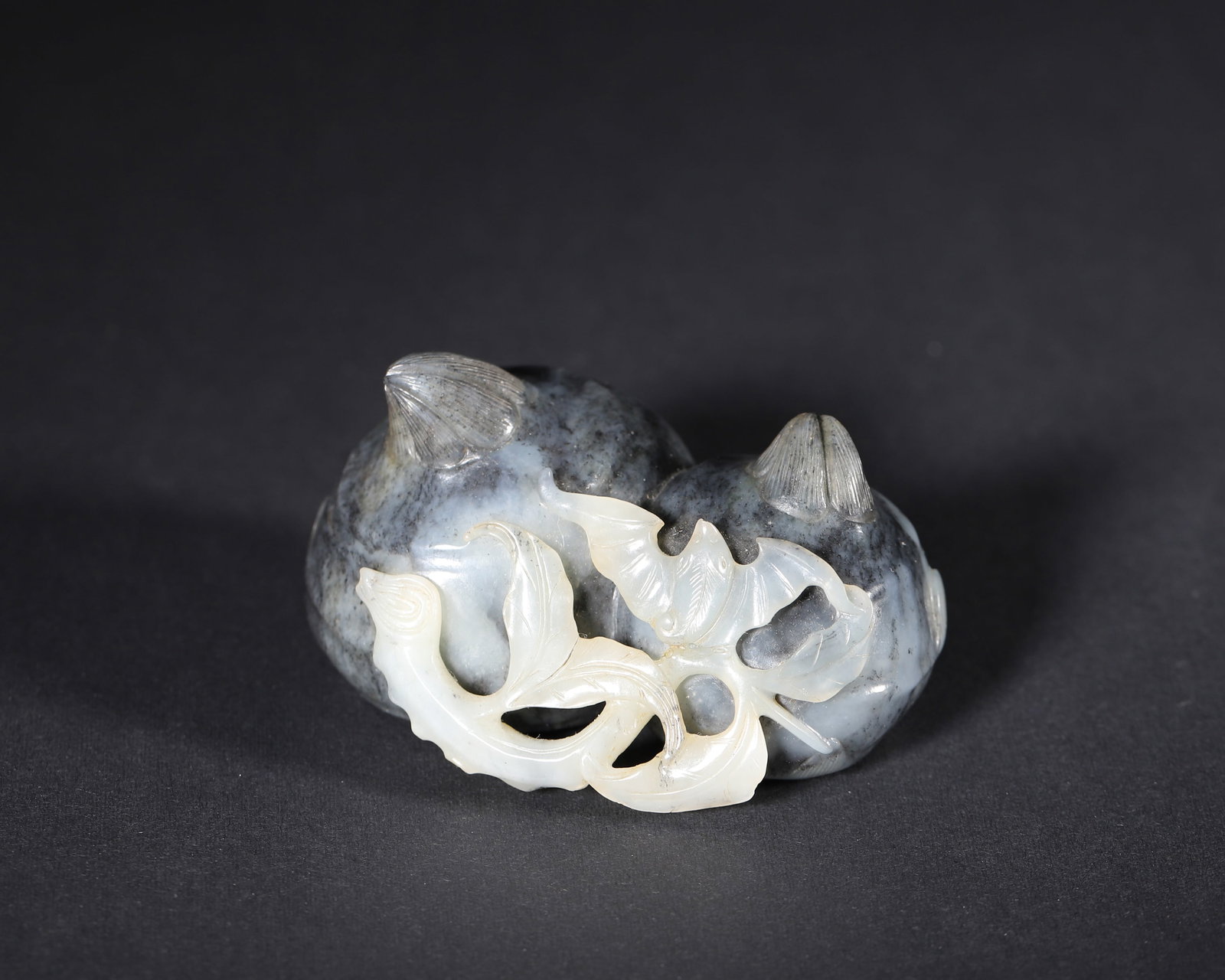 A MOTTLED JADE 'POMEGRANATE AND BAT' ORNAMENT: A mottled jade ornament carved with pomegranates, a bat and foliage, with contrasting areas of color. L. 7 cm; W. 5 cm; Ht. 4 cm; Wt. 167 g.