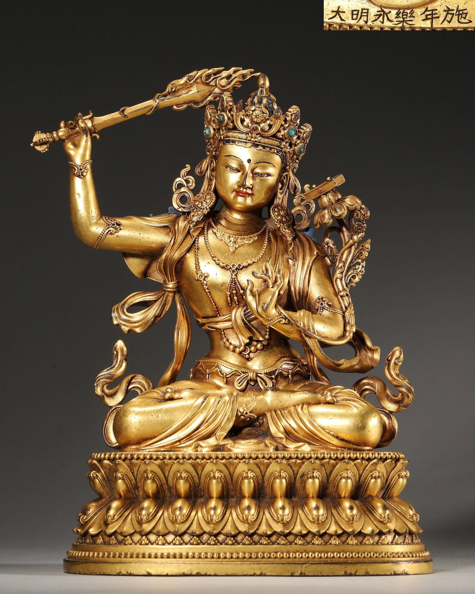 A GILT-BRONZE FIGURE OF MANJUSHRI: A gilt-bronze figure of Manjushri seated on a lotus base, holding a sword and wearing a crown set with colored stones. The base bears an inscription reading 'Da Ming Yongle Nian Shi'. L. 14 cm; W. 10