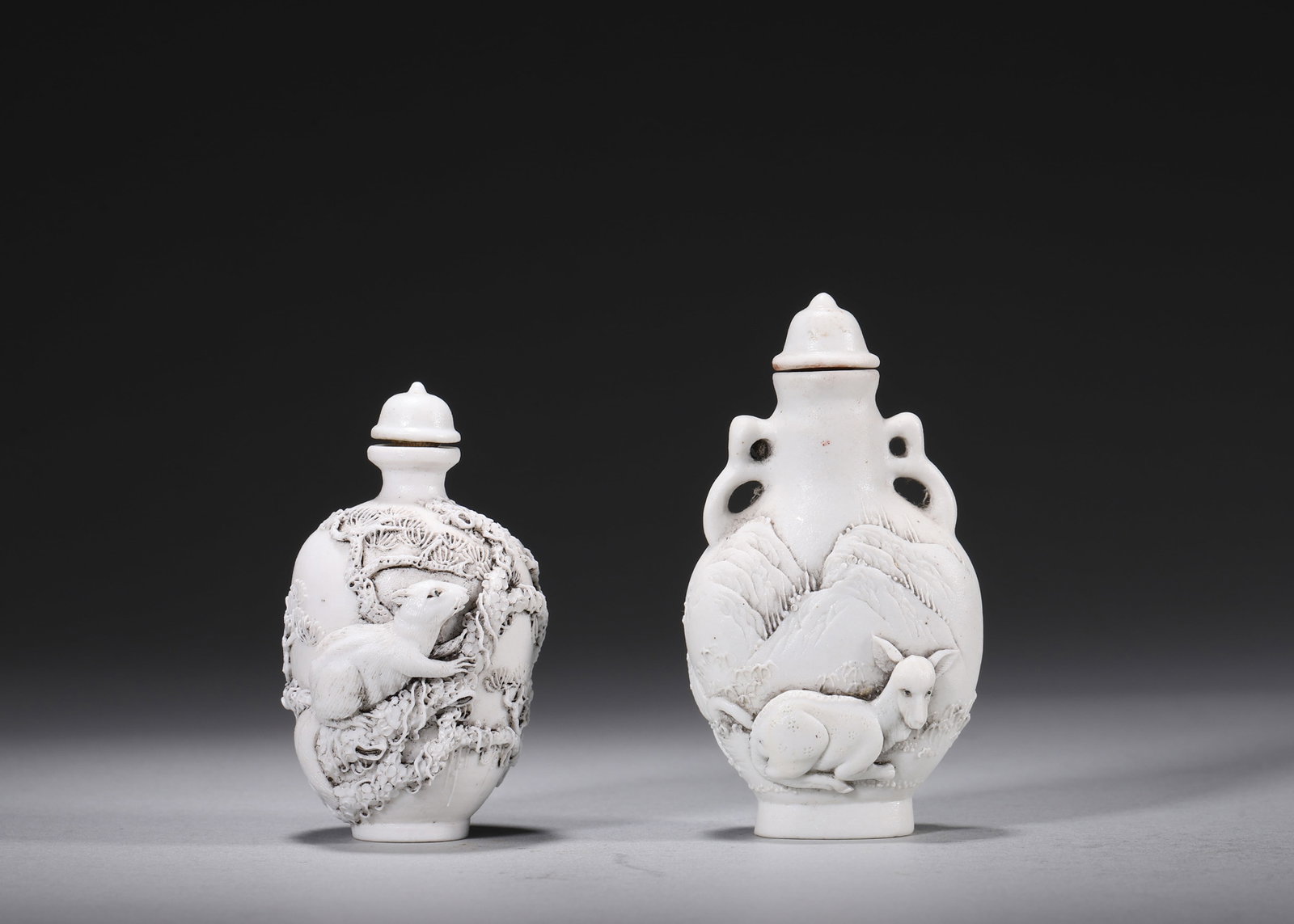 A PAIR OF WHITE-GLAZED RELIEF 'RAT AND DEER' PORCELAIN SNUFF BOTTLES: A pair of white-glazed porcelain snuff bottles carved in relief, one with a rat among pine branches and the other with a resting deer in a mountain landscape, with porcelain covers. The bases bear sea