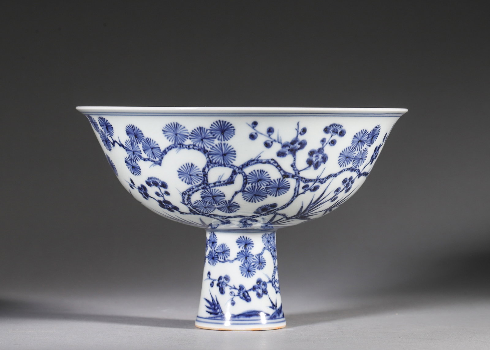 A BLUE-AND-WHITE 'PINE, BAMBOO AND PLUM' STEM BOWL: A blue-and-white porcelain stem bowl painted with pine, bamboo and plum blossoms, with floral decoration to the stem. The interior bears a six-character mark described in the source as Xuande. Diam.