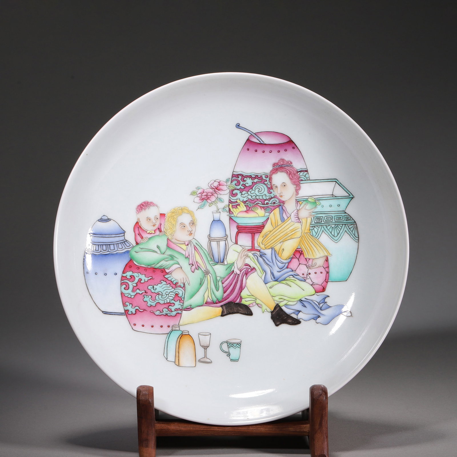 A ROUGE-RED-GROUND FAMILLE-ROSE 'EUROPEAN FIGURES' PLATE: A porcelain plate with a rouge-red-glazed exterior and a white-ground famille-rose interior painted with European figures, antiquities, fruit, peonies and Western-style vessels. The base bears a six-c