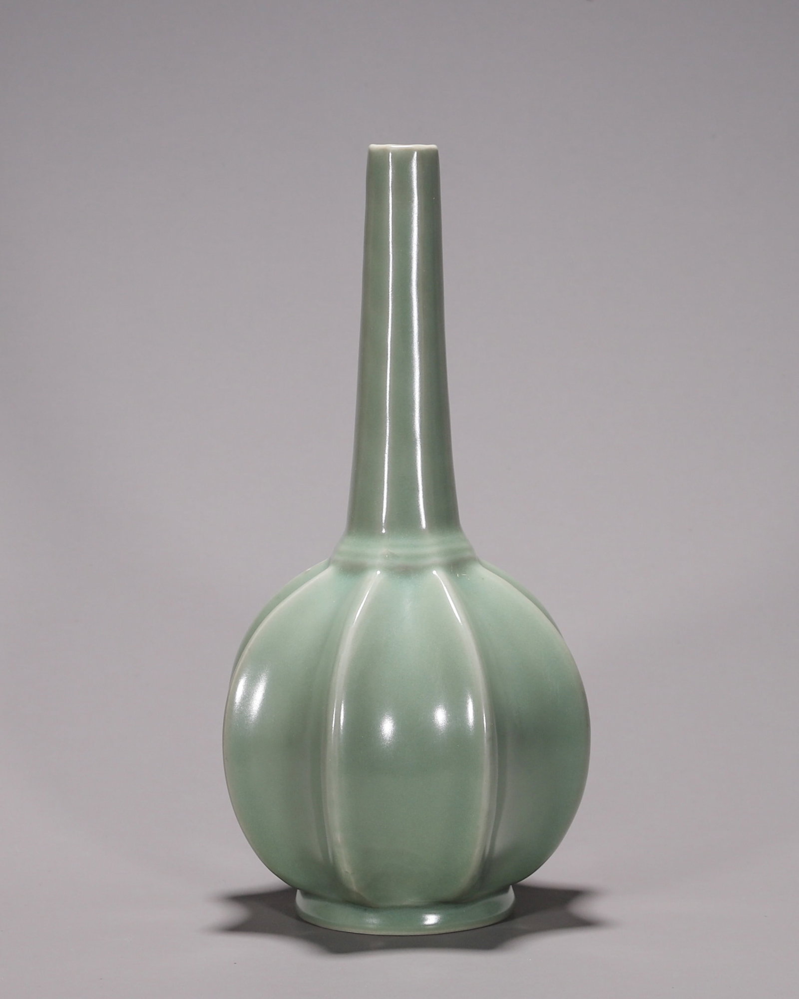 A LONGQUAN-TYPE CELADON MELON-FORM LONG-NECK VASE: A porcelain vase with a melon-ribbed body and slender neck, covered in a pinkish celadon glaze, with kiln-support marks to the foot. Diam. 13 cm; Ht. 27.5 cm.