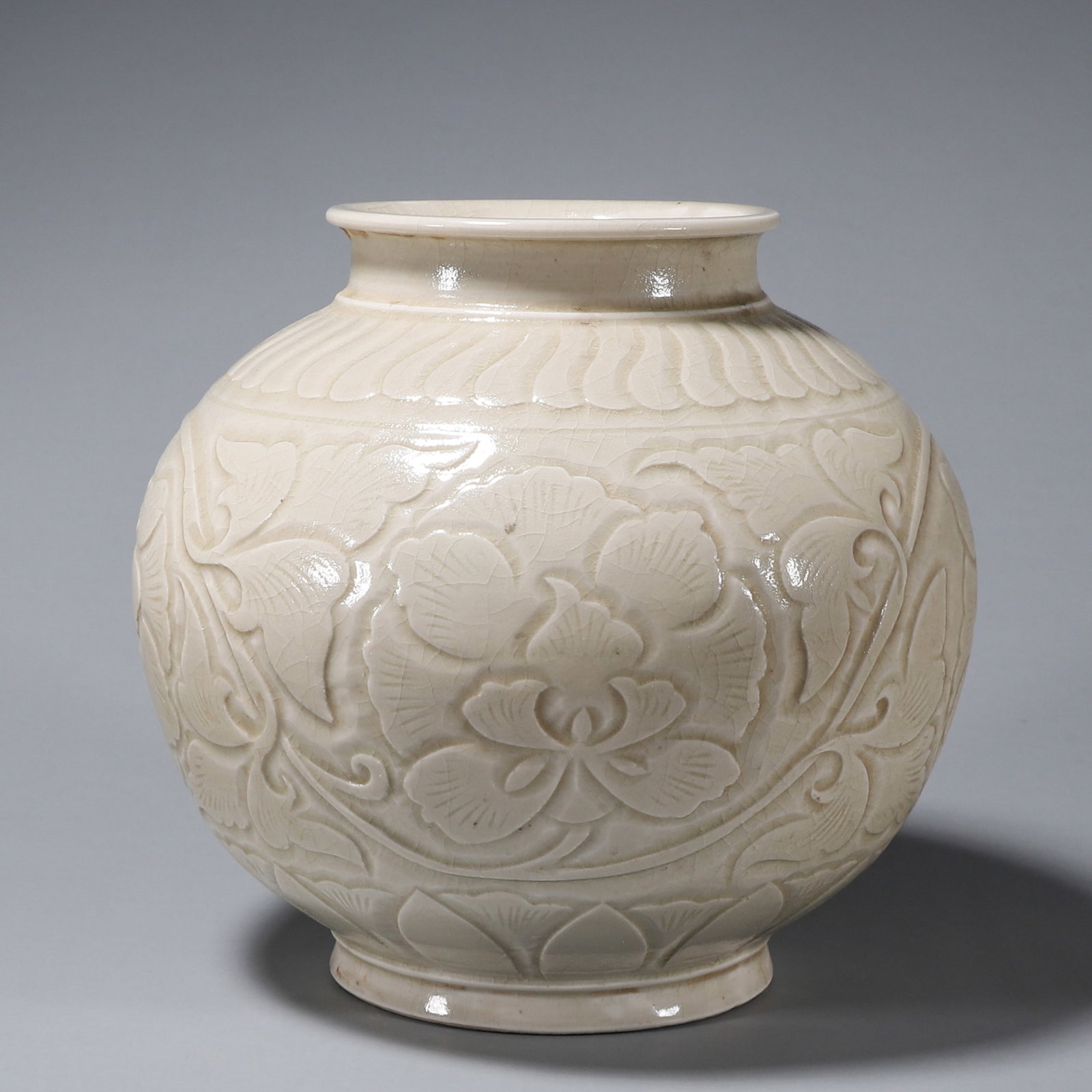 A WHITE-GLAZED CARVED 'PEONY SCROLL' PORCELAIN JAR: A porcelain jar covered in a white crackle glaze, carved with chrysanthemum-petal motifs to the shoulder and raised peony scrolls to the body, on a ring foot. Diam. 16 cm; Ht. 18 cm; Wt. 950 g.