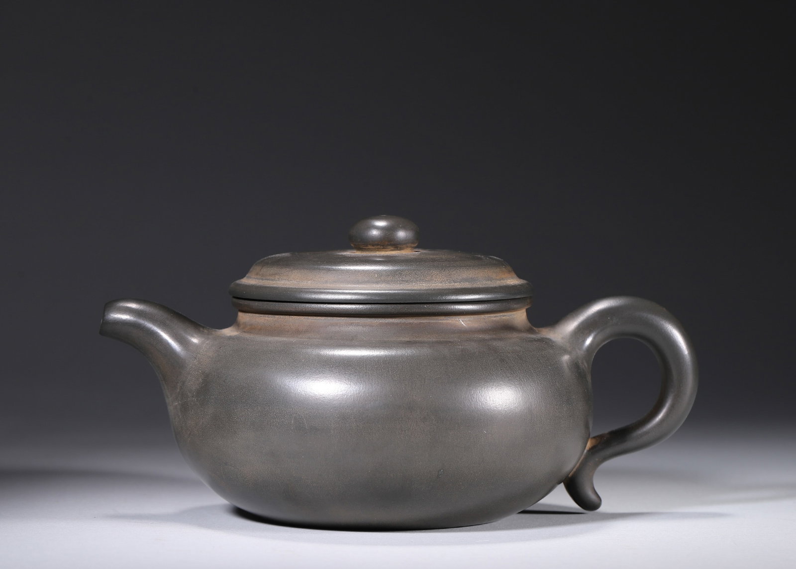 A ZISHA ARCHAISTIC TEAPOT: A dark zisha teapot with a rounded body, short spout, curved handle and bead-form finial. The base bears a seal in the name of Gu Jingzhou. L. 20 cm; W. 15 cm; Ht. 10 cm.