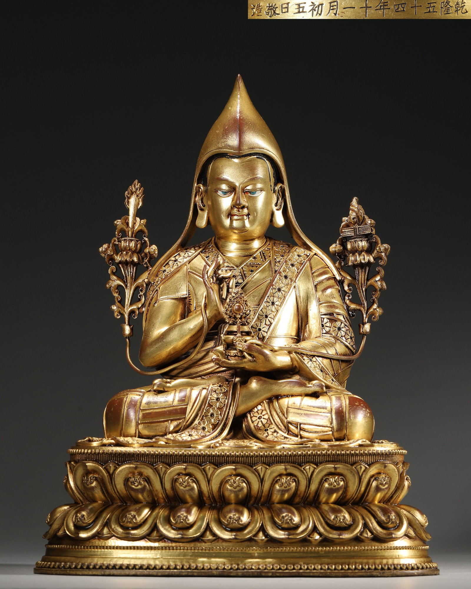 A GILT-BRONZE SEATED LAMA FIGURE: A gilt-bronze seated lama figure wearing monastic robes and holding ritual implements, on a lotus base. The figure bears a dated inscription reading 'Qianlong 54th Year, 11th Month, 5th Day, Jing