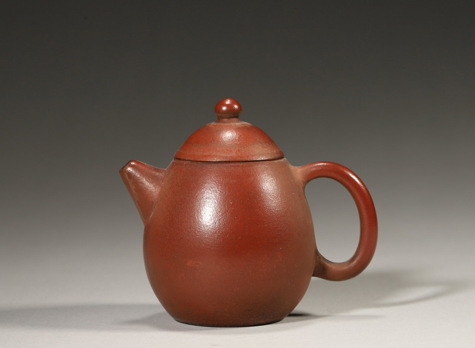 A ZISHA LONGDAN-FORM TEAPOT: A red zisha teapot of ovoid longdan form with a domed cover, bead-form finial, short spout and loop handle. The base bears a mark. L. 8 cm; W. 5.5 cm; Ht. 7 cm.