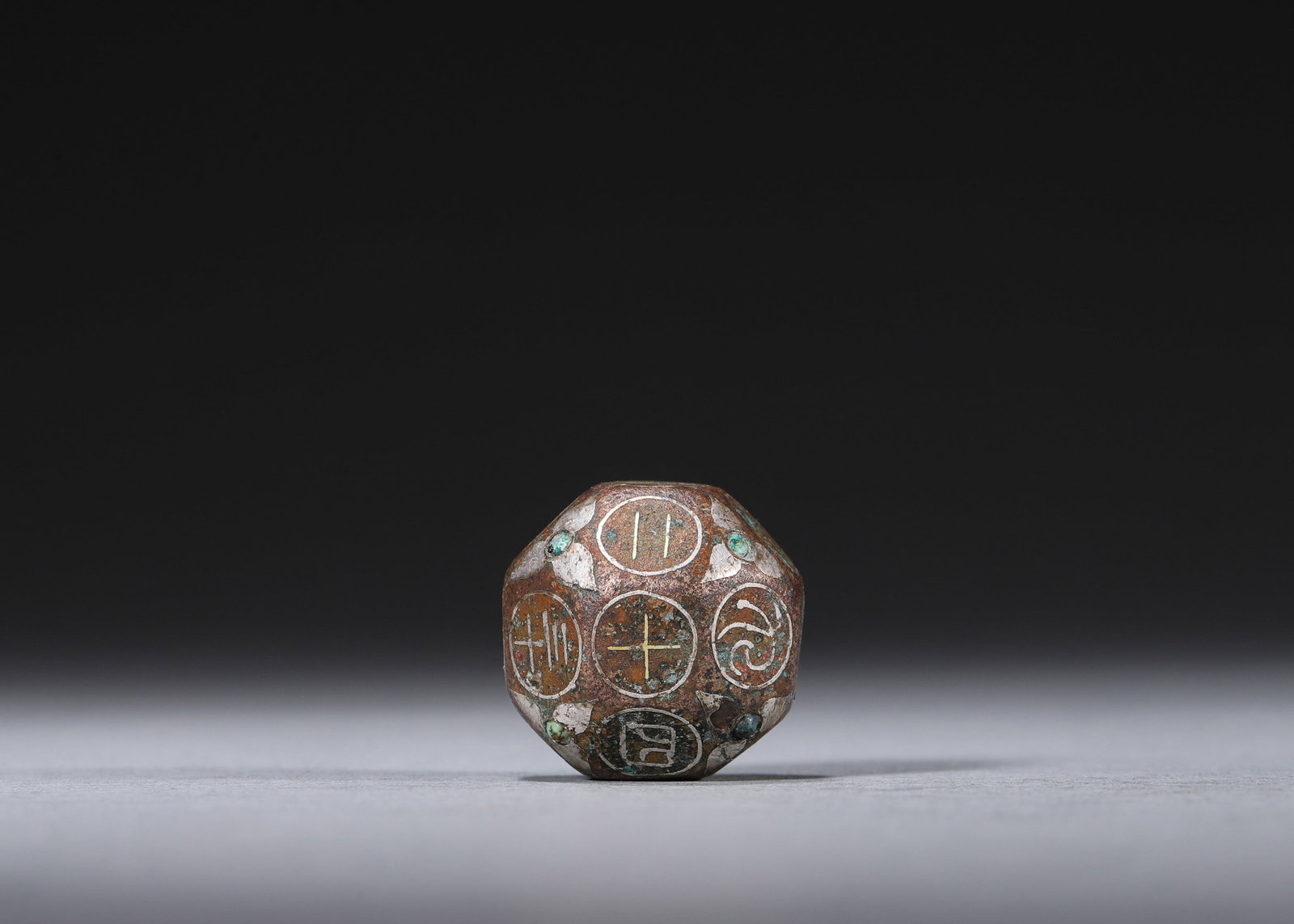 A SILVER-INLAID BRONZE 18-SIDED GAMING DIE WITH TURQUOISE INLAY: An 18-sided bronze gaming die with silver-inlaid seal-script characters and turquoise inlay to the edges. W. 2.2 cm; Wt. 50 g. Note: By repute, associated with the ancient bo game.