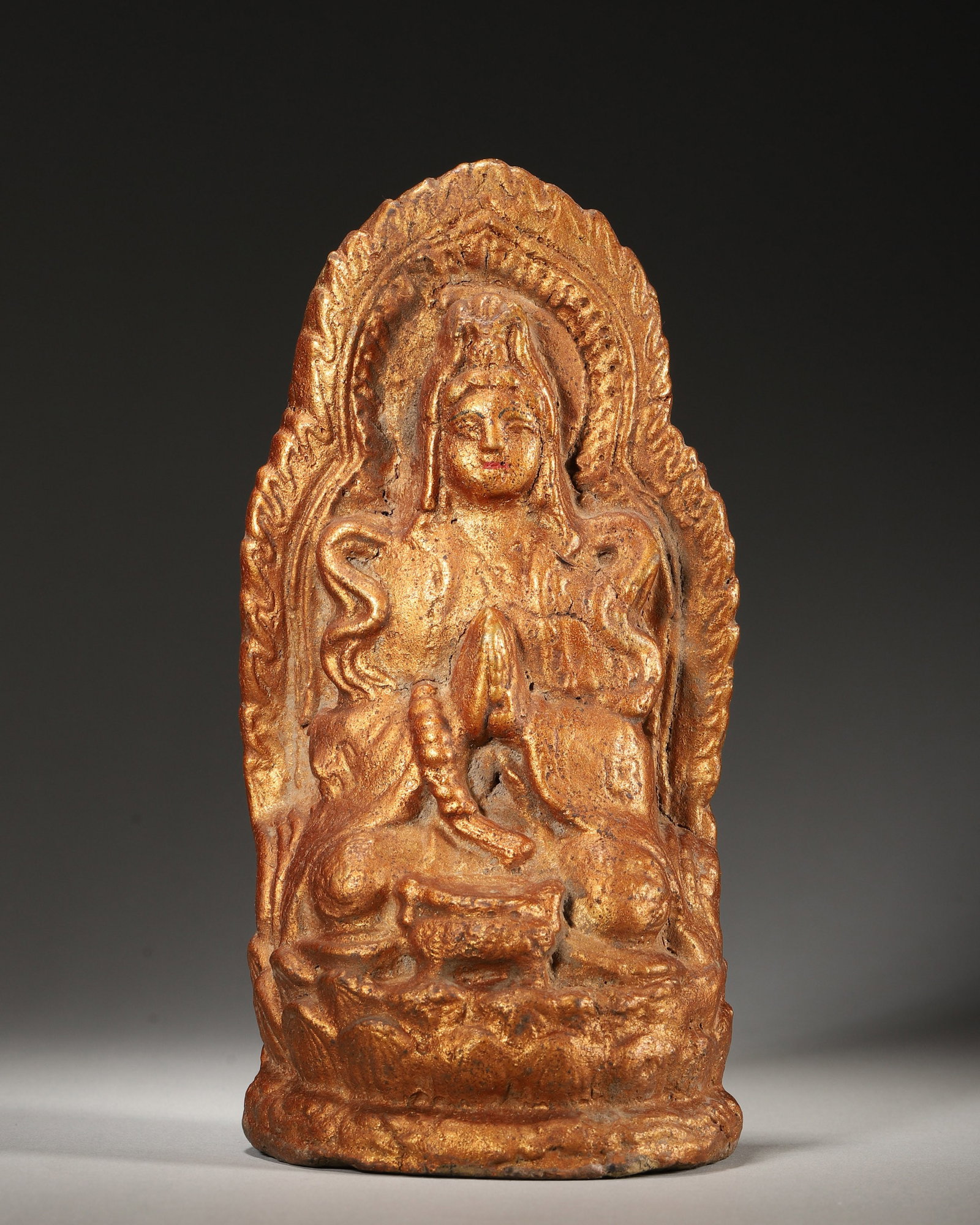 A GILT CLAY TSA-TSA OF GUANYIN: A gilt clay tsa-tsa depicting Guanyin seated on a lotus base before a flame-form mandorla. The base bears an ink inscription. W. 9.5 cm; Ht. 19 cm; Wt. 345 g. Note: By repute, associated with Tibetan