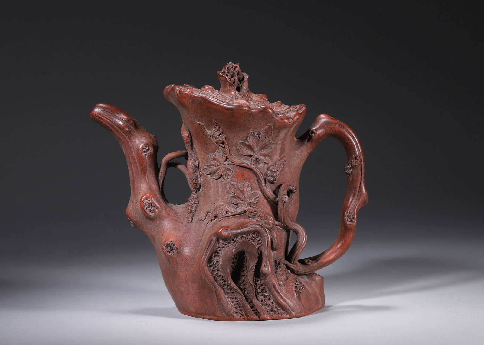 A ZISHA TREE-TRUNK-FORM TEAPOT WITH GRAPE VINES: A purple zisha teapot modeled as an old tree trunk, decorated in relief with grape vines and leaves, with branch-form spout and handle. The base and inner lid bear seals in the name of Zhu Kexin. L.