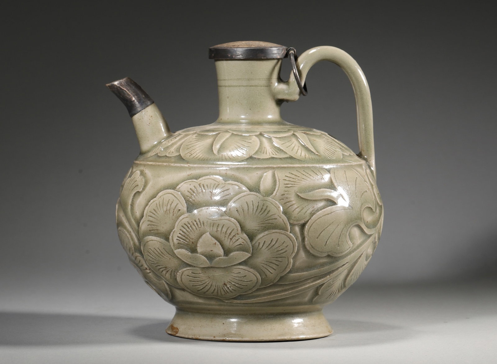 A YAOZHOU-TYPE CELADON-GLAZED CARVED FLORAL EWER: A porcelain ewer with straight neck, curved spout and handle, covered in a celadon glaze and carved with scrolling floral motifs. The foot rim is unglazed. Diam. 15 cm; Ht. 15 cm; Wt. 658 g.