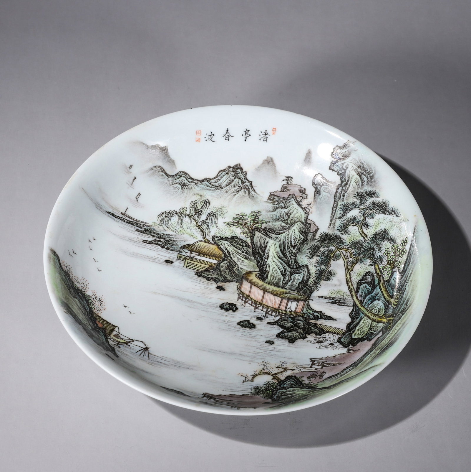 A FAMILLE-ROSE LANDSCAPE PORCELAIN PLATE: A famille-rose porcelain plate painted with a landscape and pavilion and bearing an inscription described in the source as 'Spring Waves at Riverside Pavilion'. Diam. 20.3 cm; Ht. 4.3 cm; Wt. 340 g.