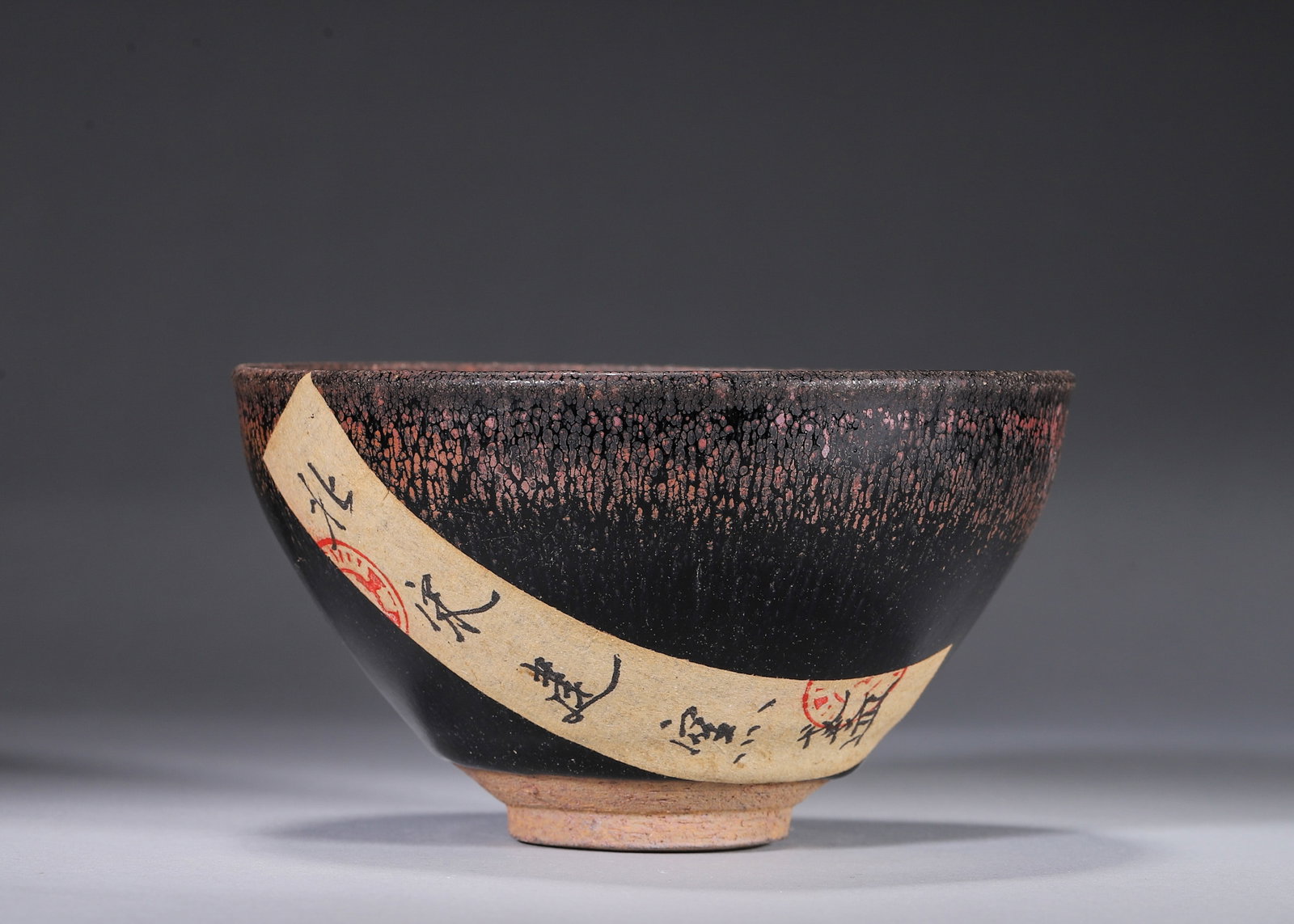 A JIAN-STYLE RED OIL-SPOT TEA BOWL: A deep tea bowl covered in a glossy black glaze with radiating reddish-brown oil-spot markings. The base bears an incised mark. Diam. 13 cm; Ht. 7.5 cm. Note: With labels.