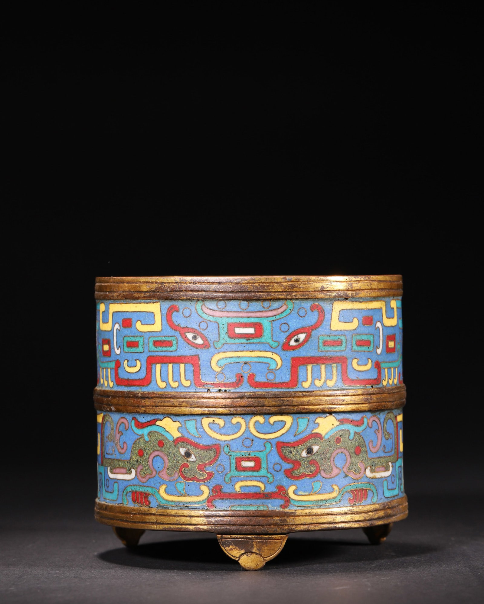 A GILT-BRONZE CLOISONNE ENAMEL TRIPOD CENSER: A cylindrical gilt-bronze tripod censer on ruyi-form feet, decorated in cloisonne enamel with archaistic beast-mask and kui-dragon motifs on a blue ground. The base bears a four-character mark describ