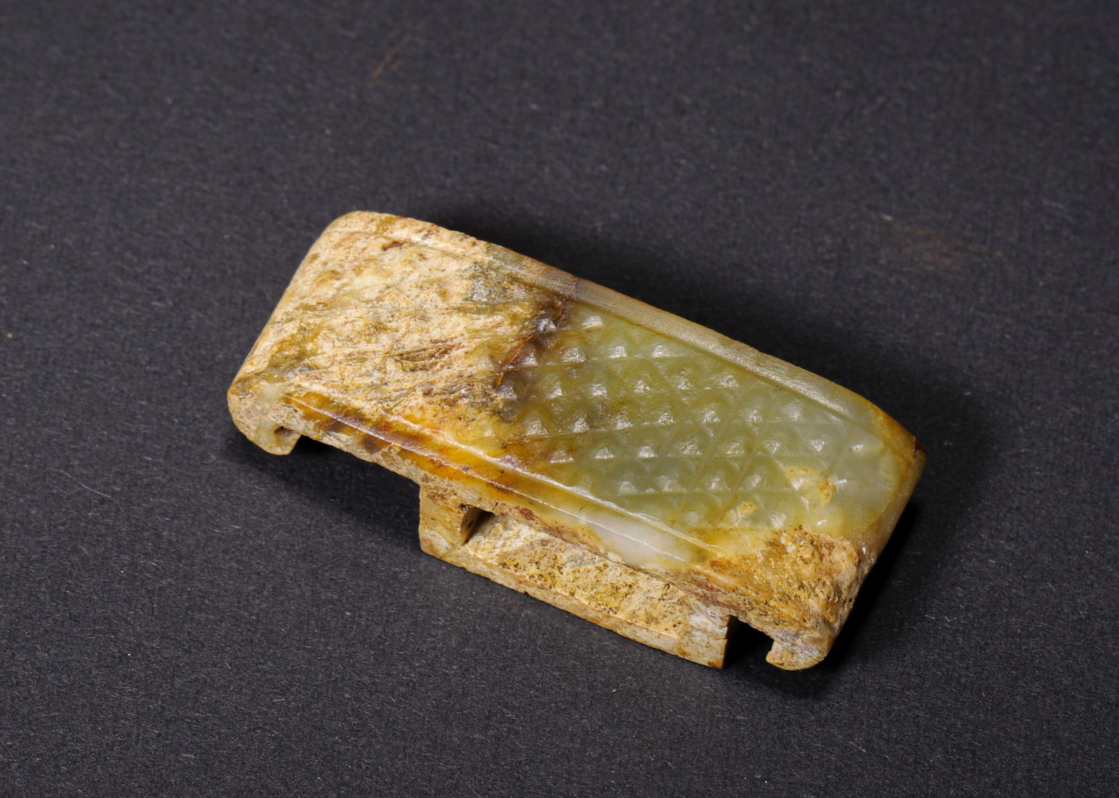 A GREEN JADE SCABBARD SLIDE WITH WOVEN-MAT DESIGN: An arched, oblong green jade scabbard slide with inward-curved ends, decorated with a woven-mat pattern, with a rectangular opening on the underside and yellow-brown surface areas. L. 4.3 cm (1.69 in)
