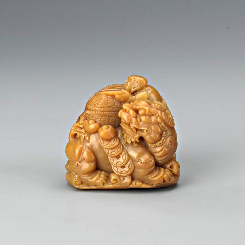 Chinese Carved Soapstone Seal: Powerfully rendered as a recumbent Piyao, carrying a string of ancient coins and a man with woven basket, his paws, mane and tail finely rendered from the yellowish tone stone with elegant details. He