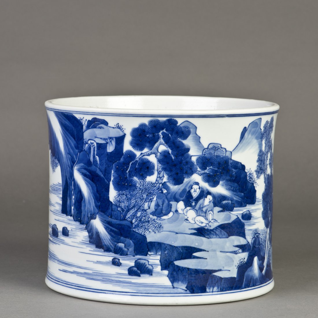 A BLUE AND WHITE PORCELAIN BRUSH POT: The classic porcelain brush pot featuring cobalt decoration of landscape with pine trees and rocks, with a guy seated on top, a boy standing on a canoe ink the lake, two figures walking and conversing