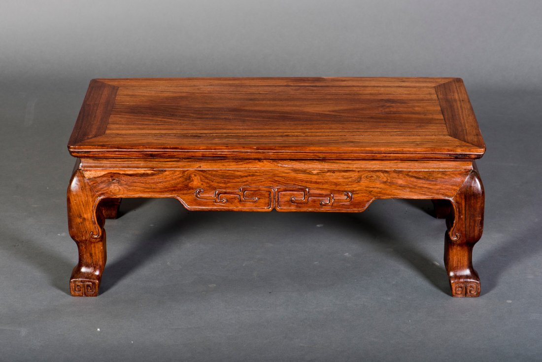 A SMALL RECTANGULAR HUANGHUALI KANG TABLE: The paneled top is set within the rectangular frame above a narrow waist and beaded, cusped aprons carved with confronting ruyi scrolls, and raised on square-section legs carved at the corners termina