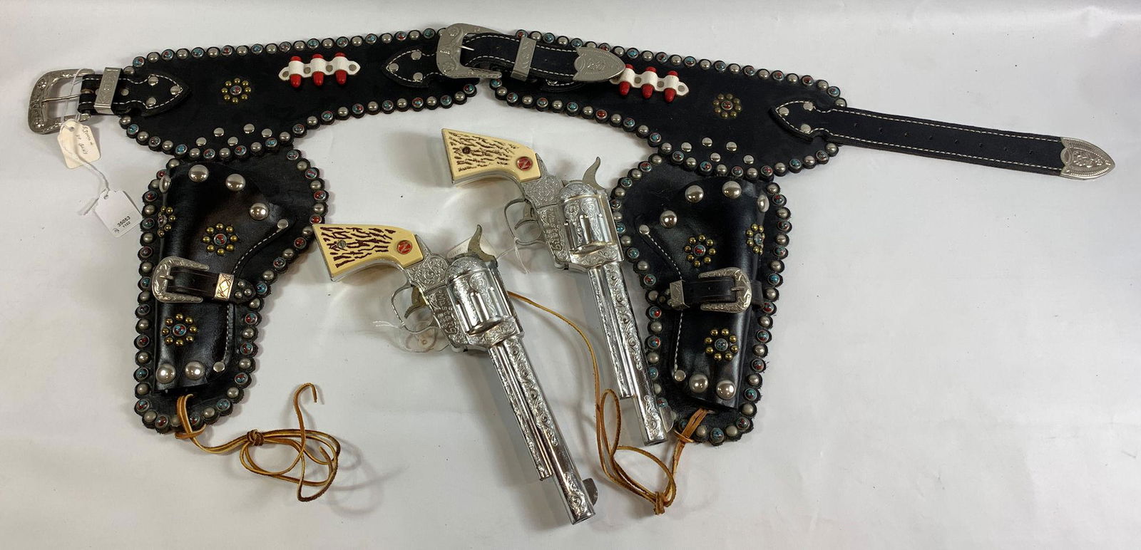 BLACK LEATHER DOUBLE HOLSTER SET. BELT AND HOLSTER: BLACK LEATHER DOUBLE HOLSTER SET. BELT AND HOLSTER DECORATED WITH TURQUOISE AND RED SWIRL STONES, SILVER AND GOLD TONE STUDS AND PAIR OF NICHOLS "MUSTANG" 500 CAP GUNS.