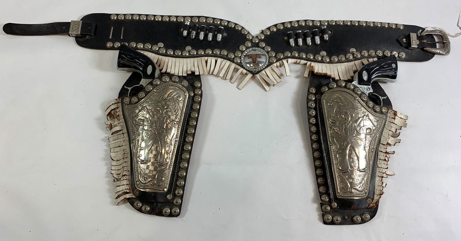 BLACK LEATHER DOUBLE HOLSTER SET, SILVER METAL STUDS: BLACK LEATHER DOUBLE HOLSTER SET, SILVER METAL STUDS AND EMBOSSED METAL FRONT WITH COWBOY, BULLETS AND FRINGE AND PAIR OF NEW BRONCO 44 CAP GUNS WITH BULLETS