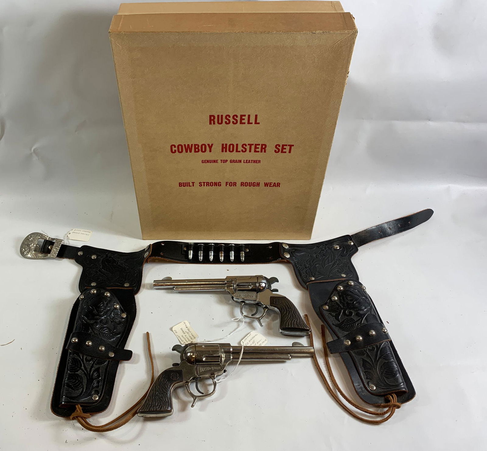 RUSSELL COWBOY HOLSTER SET IN ORIGINAL BOX INCLUDING: RUSSELL COWBOY HOLSTER SET IN ORIGINAL BOX INCLUDING BLACK LEATHER DOUBLE HOLSTERS WITH 1950-1955 G SCHMIDT BB MARSHAL BUCK'N BRONC CAP GUNS WITH COPPER STAG GRIPS