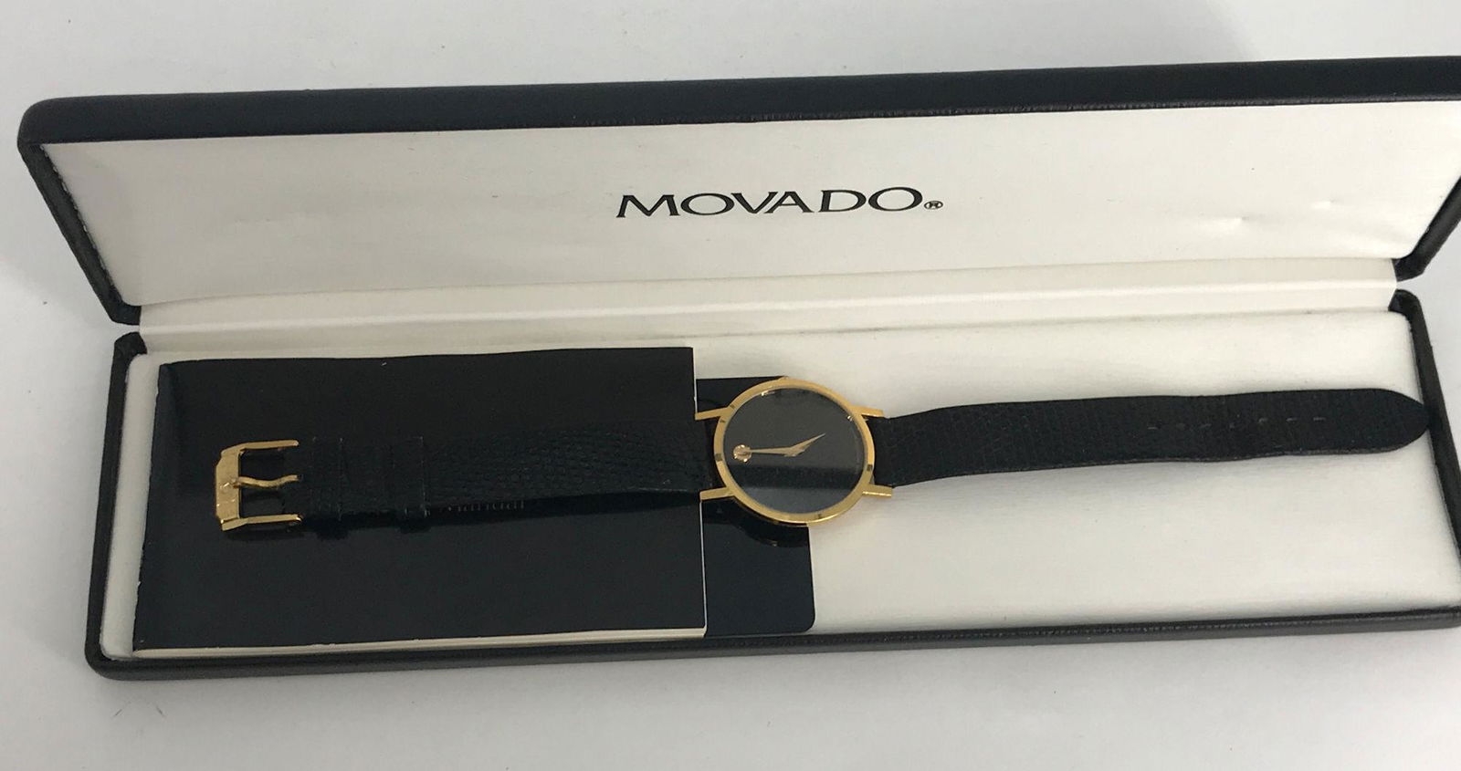 MOVADO WRIST WATCH: MOVADO WRIST WATCH ~ BLACK LEATHER BAND. GOLD TONE METAL CASE. WORN CLASP. BATTERY OPERATED QUARTZ MOVEMENT. WATER RESISTANT. IN ORIGINAL BOX WITH PAPERWORK. 9.25" IN LENGTH. NO INDICATORS. TAPERED GO