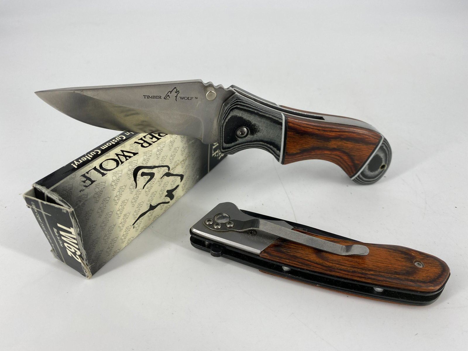 Lot of Two Folding Knives: Lot of Two Folding Knives Tac-Force folding knife with clip point blade (7.75" total length) made with 440 stainless steel. Timber Wolf knife in box numbered TW62 with clip point blade and wooden scal