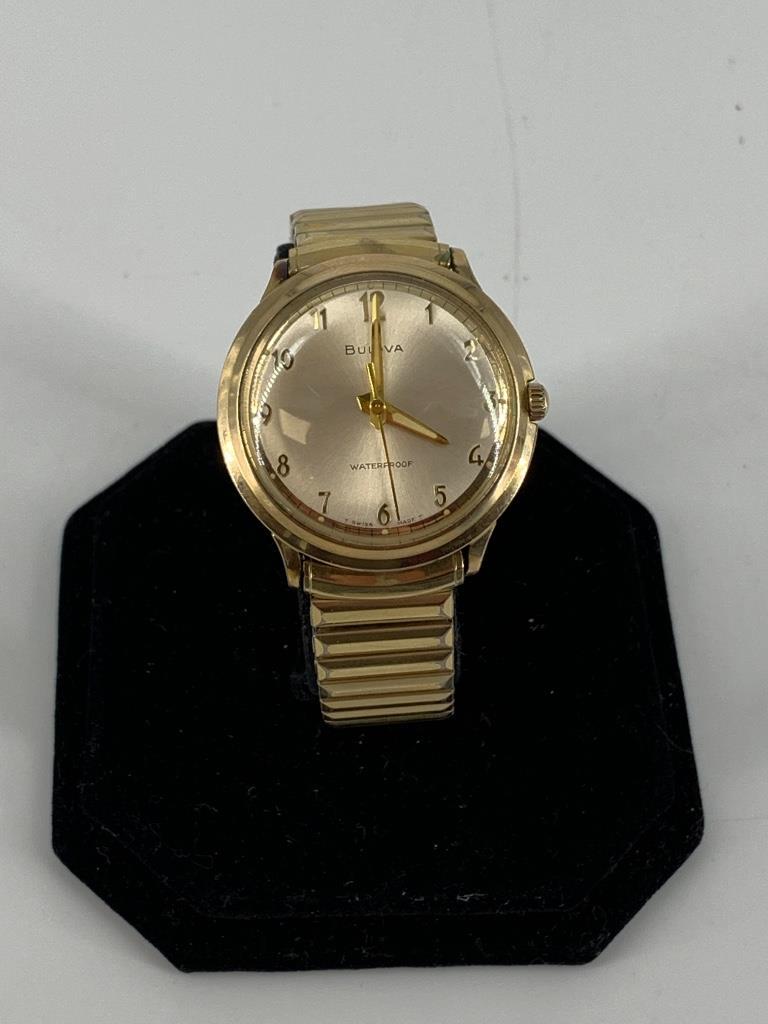 Bulova Wrist Watch: Bulova Wrist Watch Gold tone metal watch with Speidel stretch band. Off-white face with tapered gold tone hands and numeric indicators. Gold-filled case (water proof, shock resistant). Year designatio