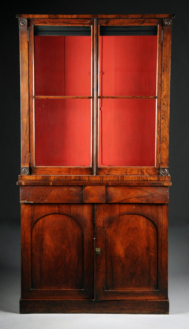 A REGENCY CARVED ROSEWOOD BOOKCASE CABINET, EARLY 19TH: A REGENCY CARVED ROSEWOOD BOOKCASE CABINET, EARLY 19TH CENTURY, the rectangular cornice above block roundel corners centering paired faux bois painted and glazed doors opening to a red painted interio