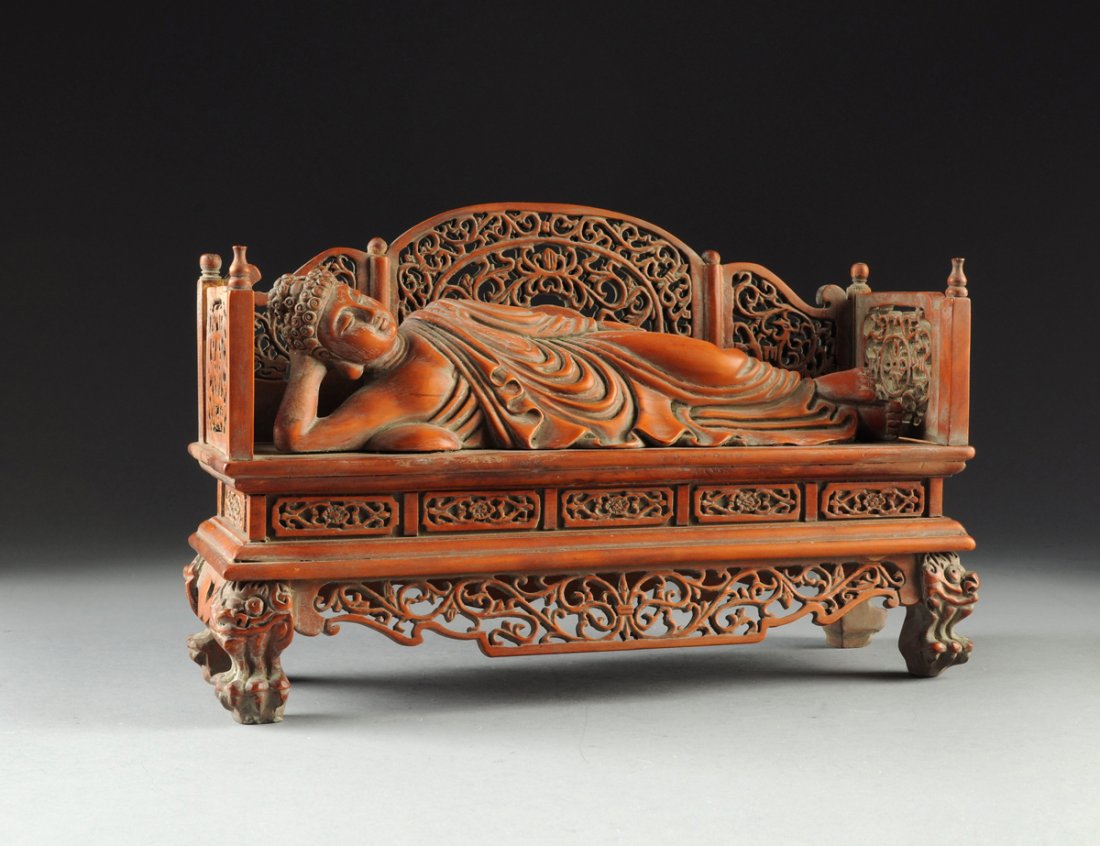 A CHINESE BOXWOOD CARVED BUDDHA RECLINING ON A BED,: A CHINESE BOXWOOD CARVED BUDDHA RECLINING ON A BED, 20TH CENTURY, the pierced carved bed frame featuring scrolls surrounding a lotus blossom, the reclining Buddha having typical tightly curled hair an
