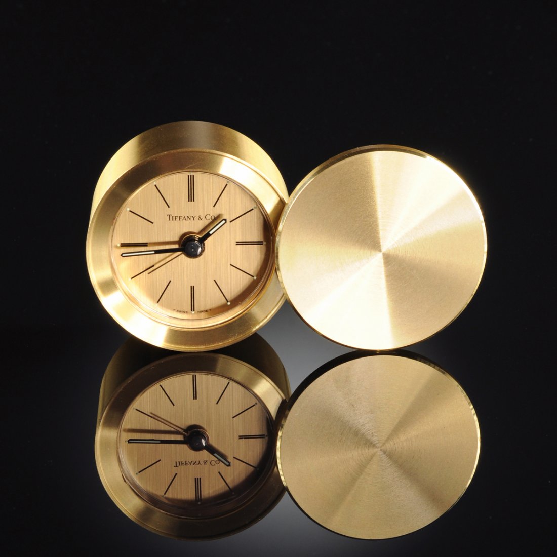 A TIFFANY & CO. GOLD TONED CIRCULAR TRAVELING CLOCK, SI: A TIFFANY & CO. GOLD TONED CIRCULAR TRAVELING CLOCK, SIGNED, THIRD QUARTER 20TH CENTURY, with conforming swivel lid above a glazed cylindrical clock centering black hour and minute chapters with sweep