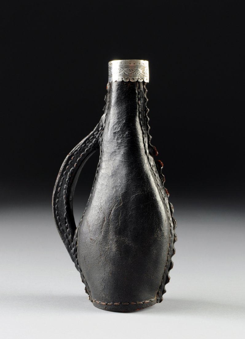 AN ANTIQUE COIN SILVER MOUNTED SEWN LEATHER GUN POWDER: AN ANTIQUE COIN SILVER MOUNTED SEWN LEATHER GUN POWDER FLASK, POSSIBLY AMERICAN, 18TH CENTURY, of flattened bottle form with one side having a serrated cut flange seam opposite a shaped flat handle an