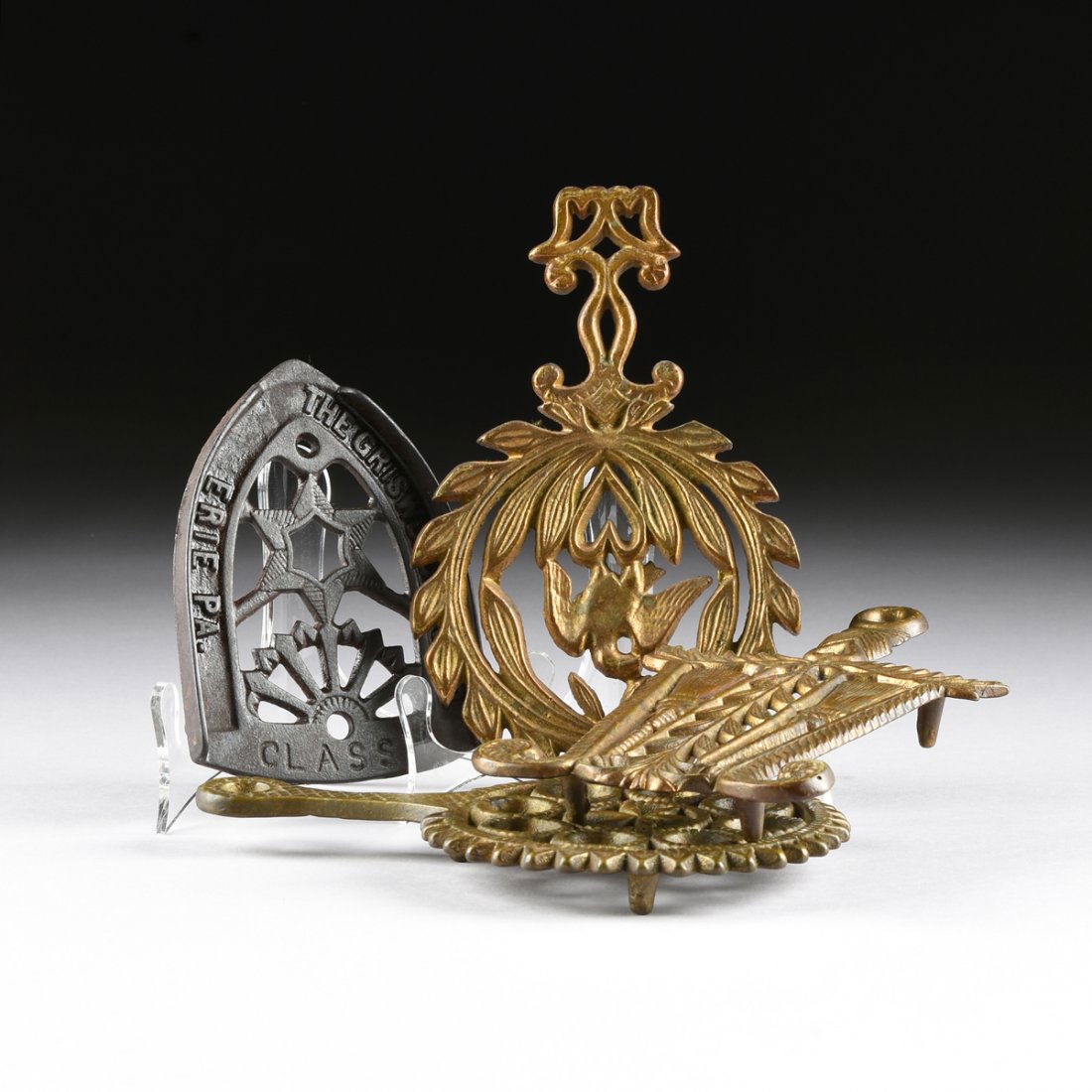 A GROUP OF FOUR AMERICAN GILT BRASS AND CAST IRON: A GROUP OF FOUR AMERICAN GILT BRASS AND CAST IRON RETICULATED TRIVETS, 19TH AND 20TH CENTURIES, comprising one with a stylized rope twist "c" scrolls handle, issuing a circular laurel leaf wreath cent