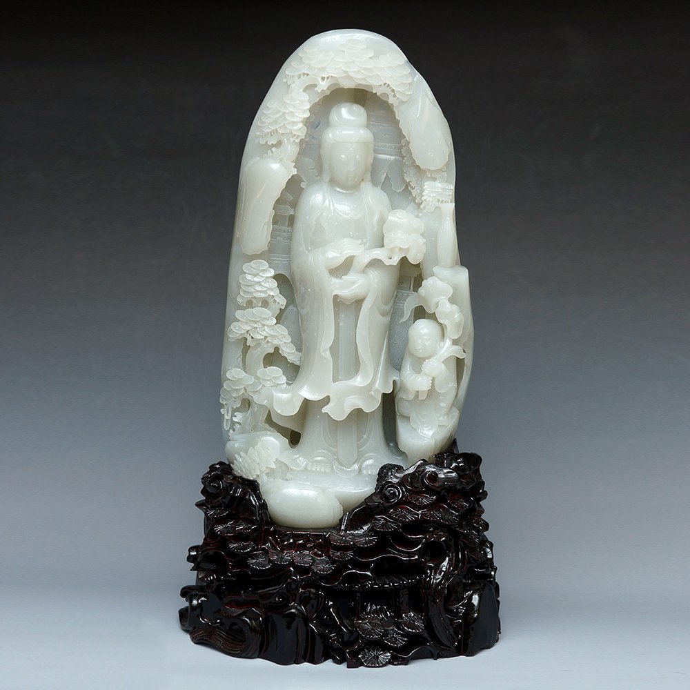 CHINESE CARVED WHITE NEPHRITE PEBBLE: Chinese carved white nephrite "pebble", the front showing guanyin and boy with pine branches overhanging; the verso with house and pine design; elaborately carved openwork wood stand. Size: 32cm x 17c