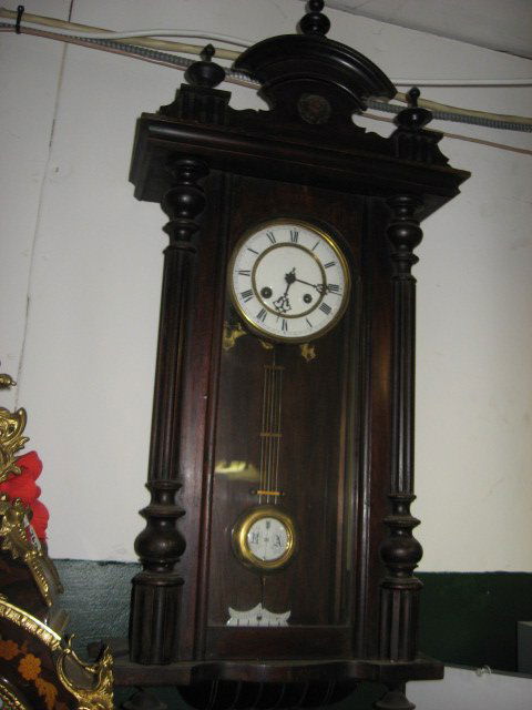 VICTORIAN-STYLE MAHOGANY WALL-HUNG CLOCK: VICTORIAN-STYLE MAHOGANY WALL-HUNG CLOCK. PORCELAIN FACE. FINIALS, COLUMNS AND ARCHES ADORN THIS CLOCK. PENDULUM HAS INITIALS "R AND A". IT HAS A SMALL DENT IN BRASS OF PENDULUM. THE TOP CENTER PORTIO