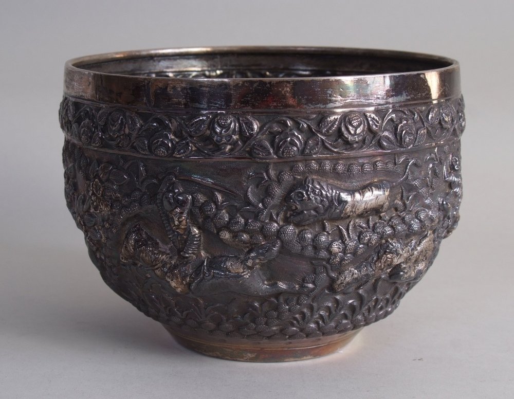 A late 19th century Burmese silver repoussé bowl, the: A late 19th century Burmese silver repoussé bowl, the tapering body depicting several hunting scenes of tiger, elephant, mounted riders, and deer, each separated with foliate-scroll, the rim with flo