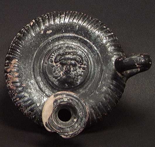GREEK CALENE WARE GUTTOS ZEUS OR HERCULES: GREEK CALENE WARE GUTTOS OIL LAMP FILLER WITH ZEUS OR HERCULES, c. 3rd century BC. The black glazed lamp filler with a facing head of Zeus or Hercules on the discus. Chip and small restored area on th