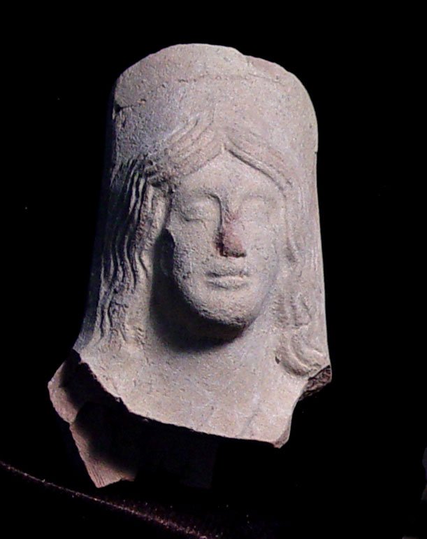 ARCHAIC GREEK VOTIVE GODDESS HEAD: AN ARCHAIC GREEK VOTIVE TERRA COTTA GODDESS. c. 4th century BC. The Korai style figure with long braided hair and wearing a polos on her head. 1.5 x 2.5 inches. Provenance: An extensive 20th century N