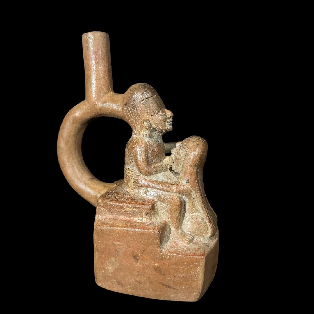 Pre-Columbian Moche Erotic pottery vessel: This remarkable pre-Columbian ceramic stirrup vessel, attributed to the Moche IV period, presents a powerful and rare depiction of a dignitary seated on a throne, receiving oral stimulation from a kne