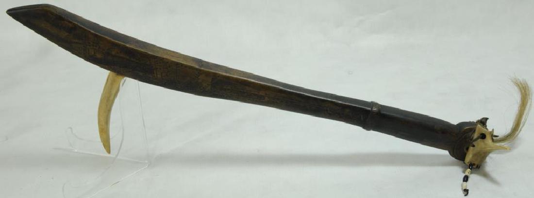 CARVED WOODEN WAR CLUB WITH LARGE TOOTH OR TUSK: PROTRUDING FROM THE END. BONE AND BEAD DECORATIONS, ORIGIN UNKNOWN. 20 1/4" H X 11 1/2" W. TUSK/TOOTH: 5" L. CLUB INCISED WITH CIRCLES, LINES, AND DOTS. END: BONE LONG NOSE MASK ATTACHED WITH HIDE. AL