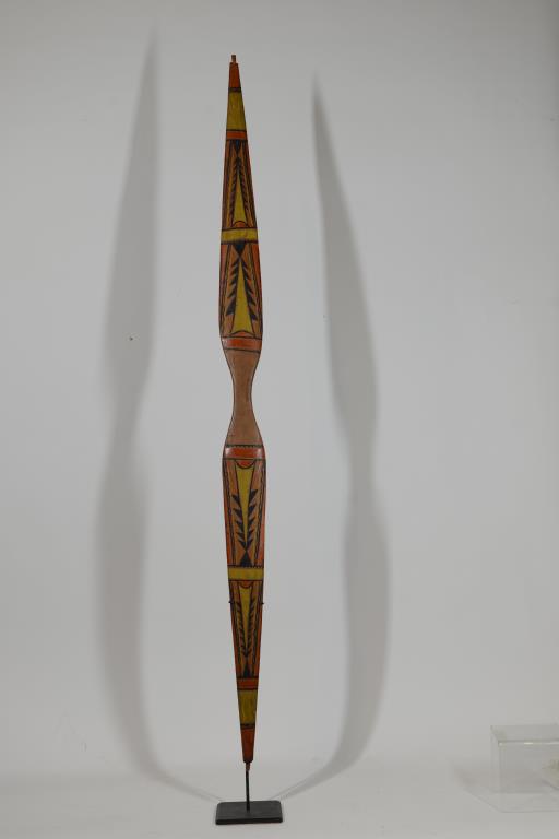 19TH C. NORTHWEST COAST CARVED WOOD AND: POLYCHROME PAINTED BOW, IN ASYMMETRICAL HOURGLASS DESIGN, WITH TWO BARBED SPEAR HEADS IN YELLOW AND BLACK, AND BRIGHT RED/ORANGE PIGMENT ON EITHER END, VERTICALLY MOUNTED ON A METAL STAND, 44" L.
