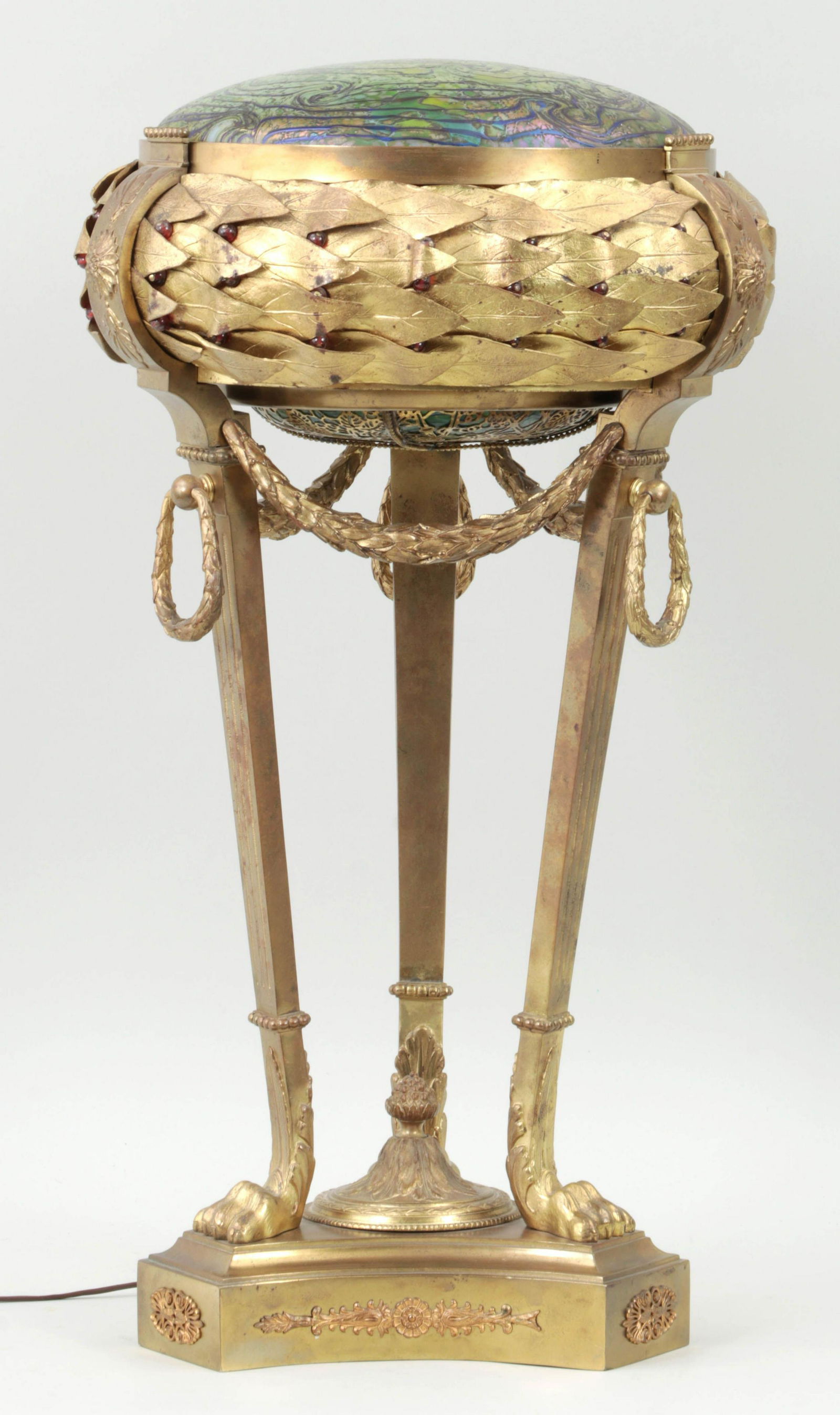 Gilt bronze Loetz table lamp: Austrian gilt bronze table lamp with Loetz iridescent glass shade and Neoclassical motifs, having round body with leaves and red bead accents, swag and swag rings, raised on fluted tripod legs ending
