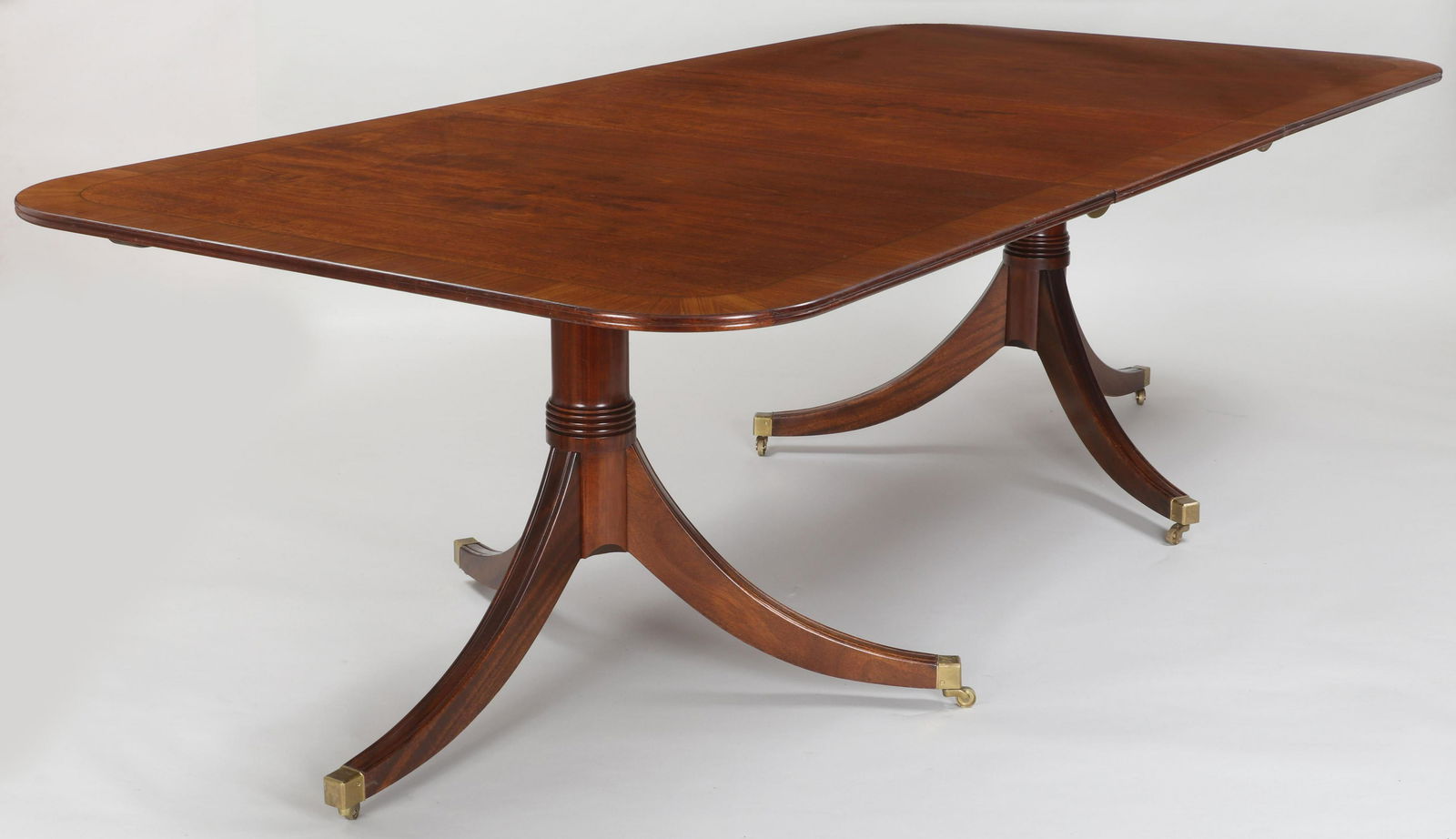 English Regency-style dining table: English Regency-style mahogany double pedestal dining table with banded top set on tripod reeded sabre legs ending on casters, having (2) leaves, 20th century. Wear to surface, minor areas of lifting
