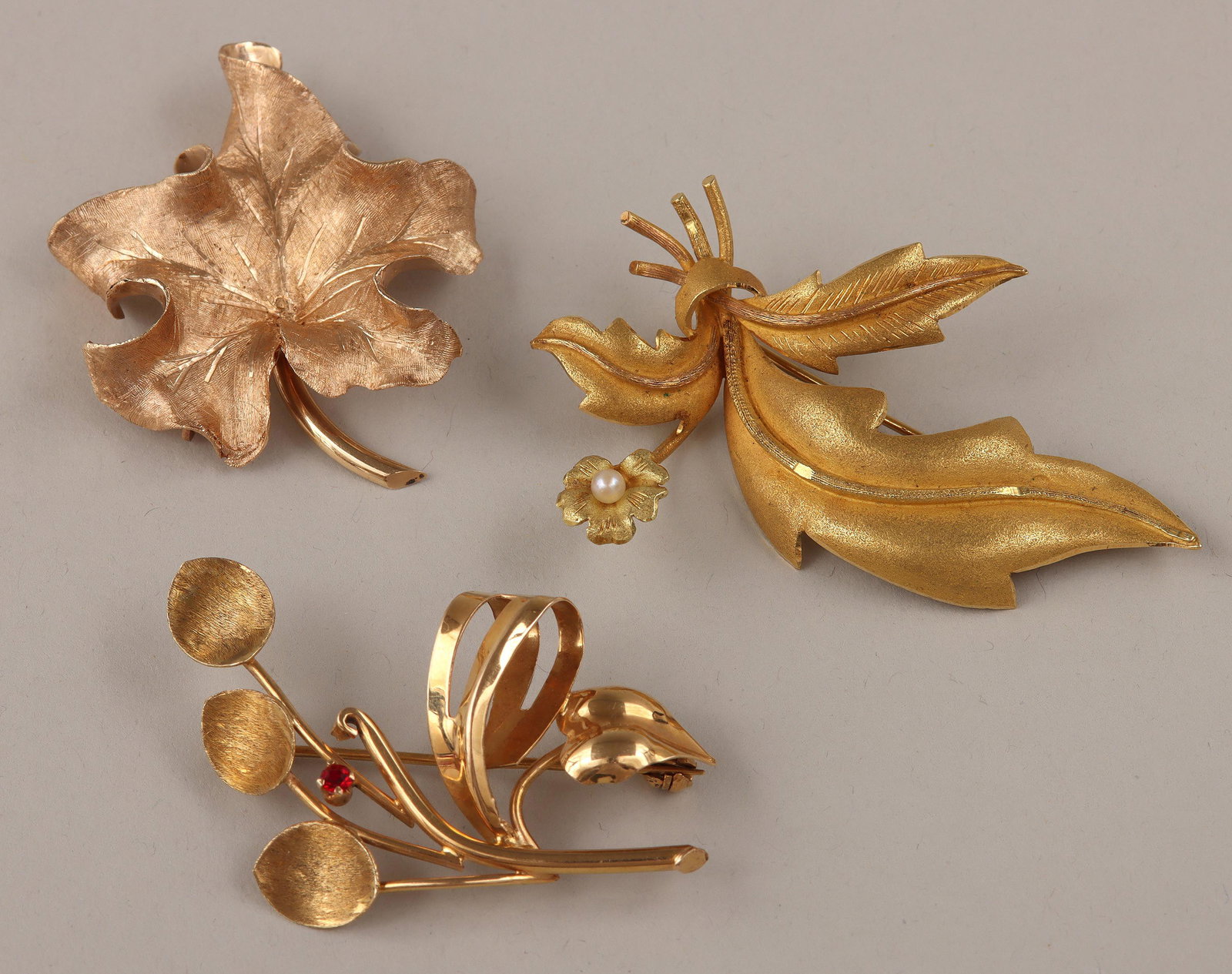 Group of (3) gold brooches: Group of (3) gold brooches, to include: 18k brooch having large leaves and small flower with seed pearl at center; 18k plant brooch, with round-cut red stone, (stone with minor nick); and 14k curled l