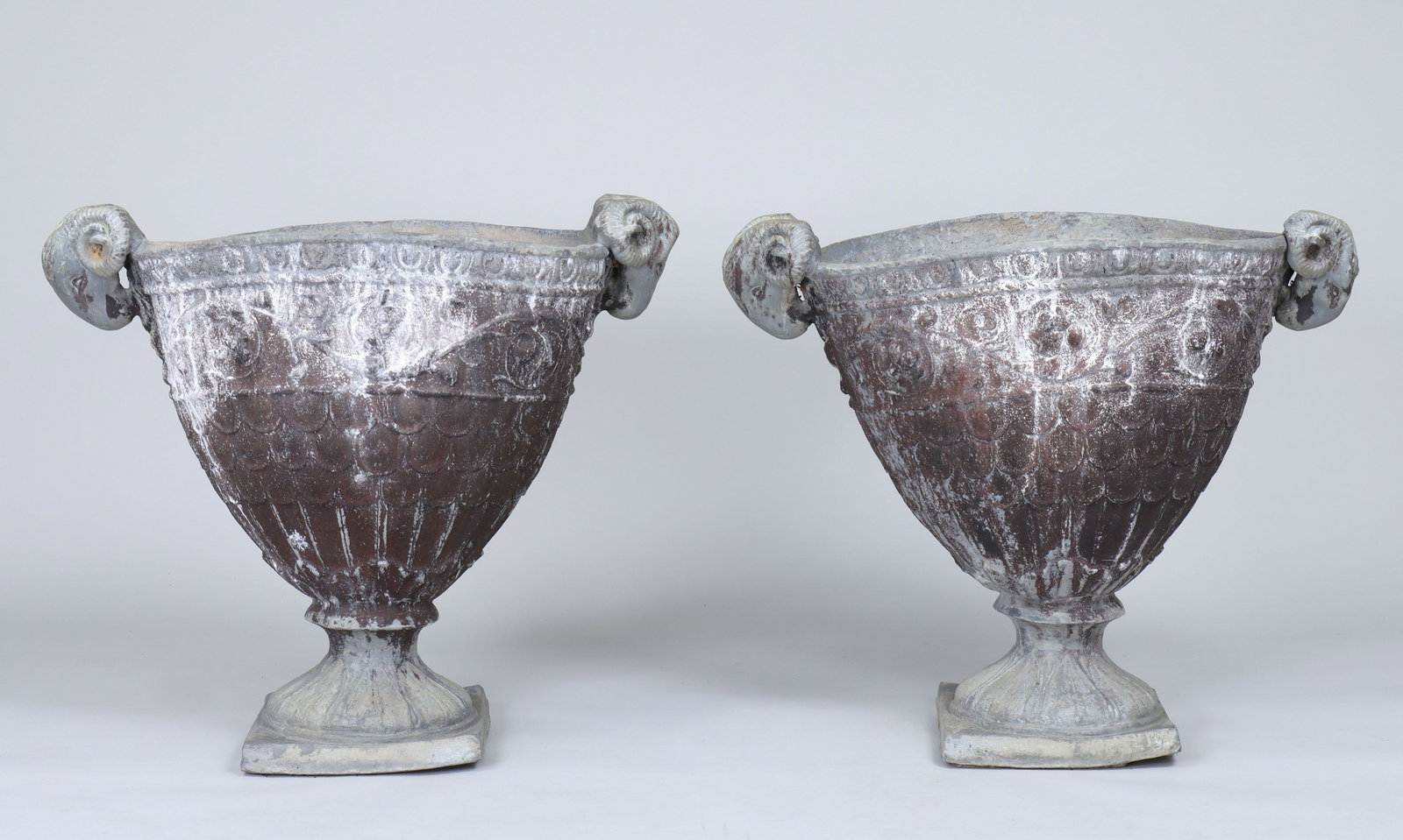 Pair of Neoclassical lead garden urns: Pair of Neoclassical lead garden urns with ram's head handles and relief-decorated bodies in foliate scrolls and scales. Misshaping, holes and repairs. 28-1/2''h, 32''w, 18-1/2''d.